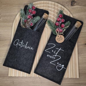 K&ouml;nnte beinhalten: Zwei dunkelgraue Filz-Geschenkkartenhalter mit wei&szlig;er Schrift. Jeder Halter enth&auml;lt ein Glasr&ouml;hrchen mit Korken, verziert mit k&uuml;nstlichen Beeren und Kiefernzweigen. Auf einem steht "Gutschein", auf dem anderen "Zeit statt Zeug".