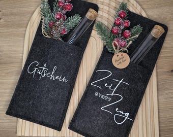 Könnte beinhalten: Zwei dunkelgraue Filz-Geschenkkartenhalter mit weißer Schrift. Jeder Halter enthält ein Glasröhrchen mit Korken, verziert mit künstlichen Beeren und Kiefernzweigen. Auf einem steht "Gutschein", auf dem anderen "Zeit statt Zeug".