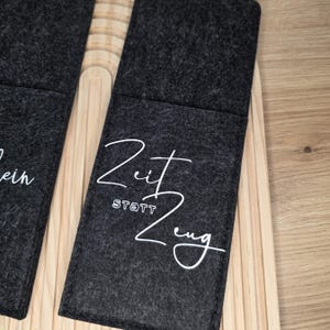 K&ouml;nnte beinhalten: Dunkelgrauer Filz-Weinflaschenhalter mit den Worten "Zeit statt Zeug" in wei&szlig;er Schrift. Der Halter ist rechteckig mit einer Tasche und vor einem Holzgrund platziert.