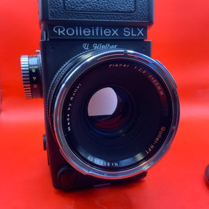 Puede incluir: Una cámara Rolleiflex SLX negra con un objetivo Planar 1:2.8 f=80mm. La cámara tiene un anillo de lente plateado y un cuerpo de cuero sintético negro. La cámara está fabricada por Rollei y tiene el número de serie Nr. 8085457.