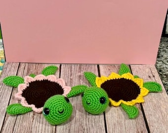 May include: Two crocheted turtle toys. One turtle has a pink and brown sunflower shell, the other has a yellow and brown sunflower shell. Both turtles have green heads, flippers, and black eyes.