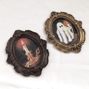 May include: Two small, oval-shaped framed pictures. One features a candle with a flame, the other a ghost holding a pumpkin. The frames are ornate, with a dark brown and gold finish, respectively. The background is a white fabric.