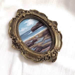 Op de afbeelding: Een ovaal schilderij van een kustlandschap met donkere rotsen, een zandstrand en een blauwe lucht met witte wolken, gevat in een sierlijke, antiek-goudkleurige lijst met gedetailleerde reli&euml;fsnijwerken langs de randen.