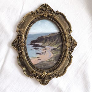 May include: An oval-shaped painting of a coastal landscape featuring a rocky cliffside meeting the ocean, encased in an ornate, antique gold-colored frame with intricate floral and shell-like relief detailing along the scalloped edges.