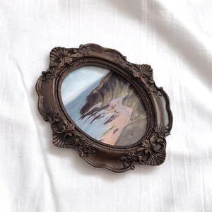 May include: An oval-shaped painting in a dark brown, ornate, decorative frame. The artwork depicts a coastal landscape with rocky cliffs, a sandy beach, and blue water. The frame features intricate relief carvings of leaves and shell-like motifs.