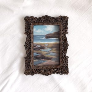 Op de afbeelding: Een rechthoekig schilderij van een kustlandschap met een rotsachtige kustlijn, blauw zeewater en een bewolkte lucht. Het kunstwerk is gevat in een sierlijke, donkerbruine, getextureerde lijst met ingewikkeld krulwerk en een parelrand.