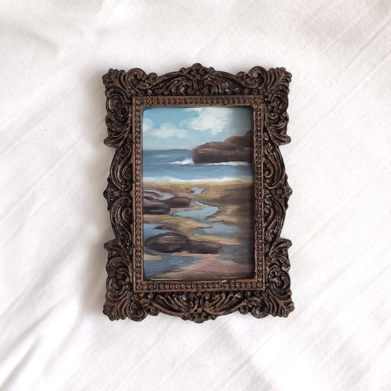 Op de afbeelding: Een rechthoekig schilderij van een kustlandschap met een rotsachtige kustlijn, blauw zeewater en een bewolkte lucht. Het kunstwerk is gevat in een sierlijke, donkerbruine, getextureerde lijst met ingewikkeld krulwerk en een parelrand.