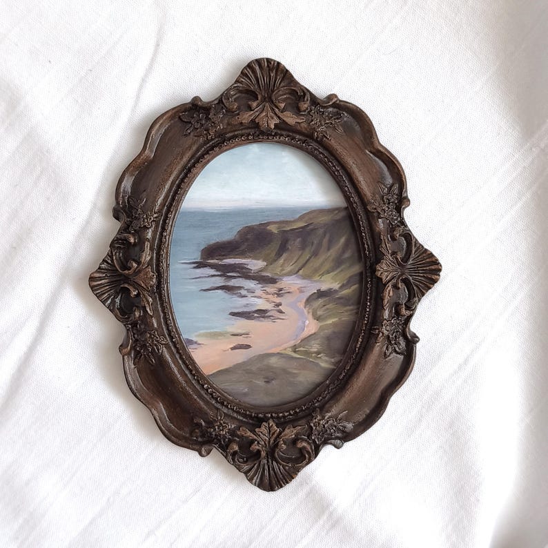 May include: An oval-shaped painting of a coastal landscape featuring a sandy beach, dark cliffs, and a blue ocean, encased in a dark brown, ornate, decorative frame with intricate floral and shell-like carvings along the outer edges.