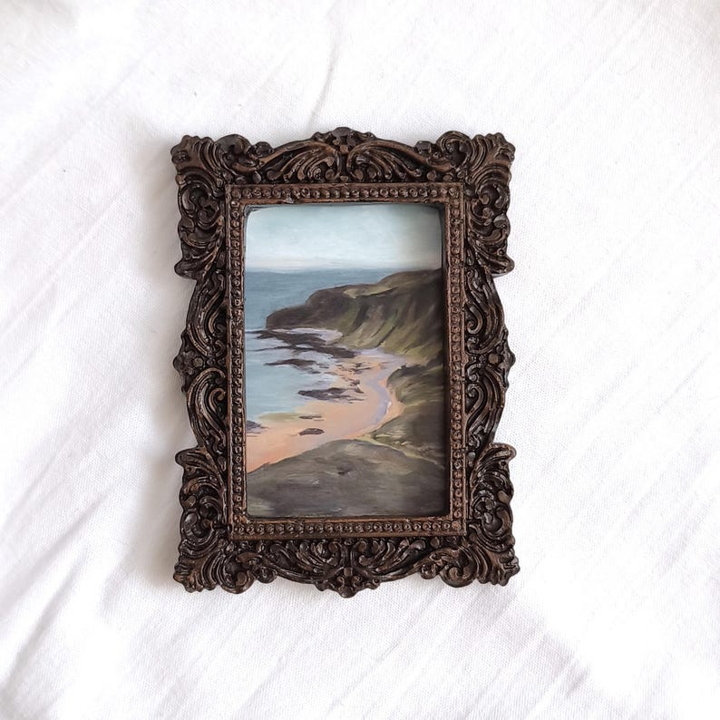 May include: A rectangular painting of a coastal landscape featuring a sandy beach, dark cliffs, and a calm sea, enclosed within an ornate, dark brown, intricately carved frame with scrollwork details and a beaded inner border edge.