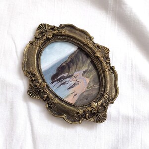 May include: An oval-shaped painting of a coastal landscape featuring cliffs and a sandy beach, encased in an ornate, antique-gold colored frame with detailed floral and shell-like relief carvings along the edges and a textured metallic finish.
