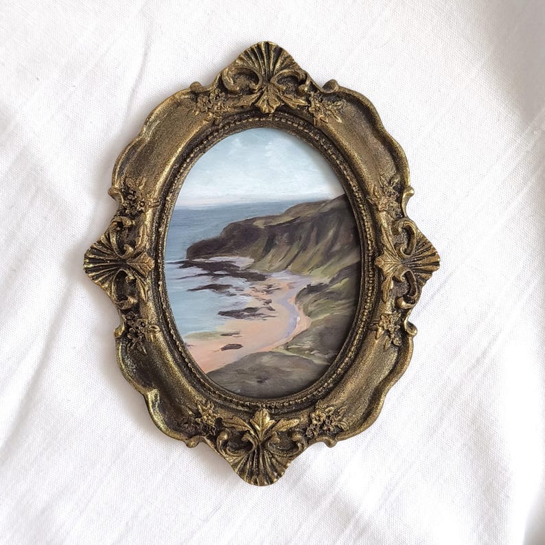 May include: An oval-shaped painting of a coastal landscape featuring a rocky cliffside meeting the ocean, encased in an ornate, antique gold-colored frame with intricate floral and shell-like relief detailing along the scalloped edges.