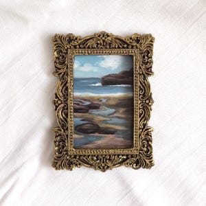 Op de afbeelding: Een rechthoekig olieverfschilderij van een kustlandschap met een rotsachtige kustlijn, blauw zeewater en een bewolkte lucht, gevat in een sierlijke, antiek ogende goudkleurige lijst met ingewikkeld krulwerk en een parelrand aan de binnenzijde.