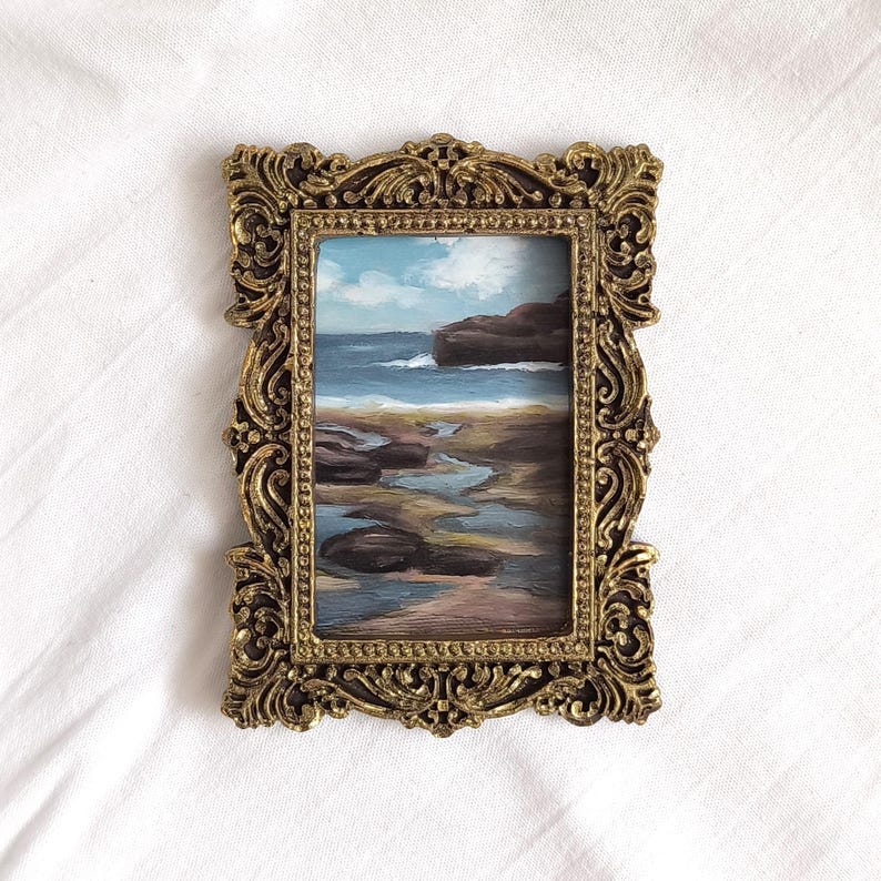 Op de afbeelding: Een rechthoekig olieverfschilderij van een kustlandschap met een rotsachtige kustlijn, blauw zeewater en een bewolkte lucht, gevat in een sierlijke, antiek ogende goudkleurige lijst met ingewikkeld krulwerk en een parelrand aan de binnenzijde.