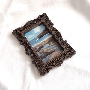 Op de afbeelding: Een rechthoekig schilderij van een kustlandschap met een blauwe oceaan, rotsachtige kustlijn en een bewolkte lucht, gevat in een sierlijke, donkerbruine, uitvoerig gesneden lijst met krulwerkdetails en een kralenrand aan de binnenzijde.