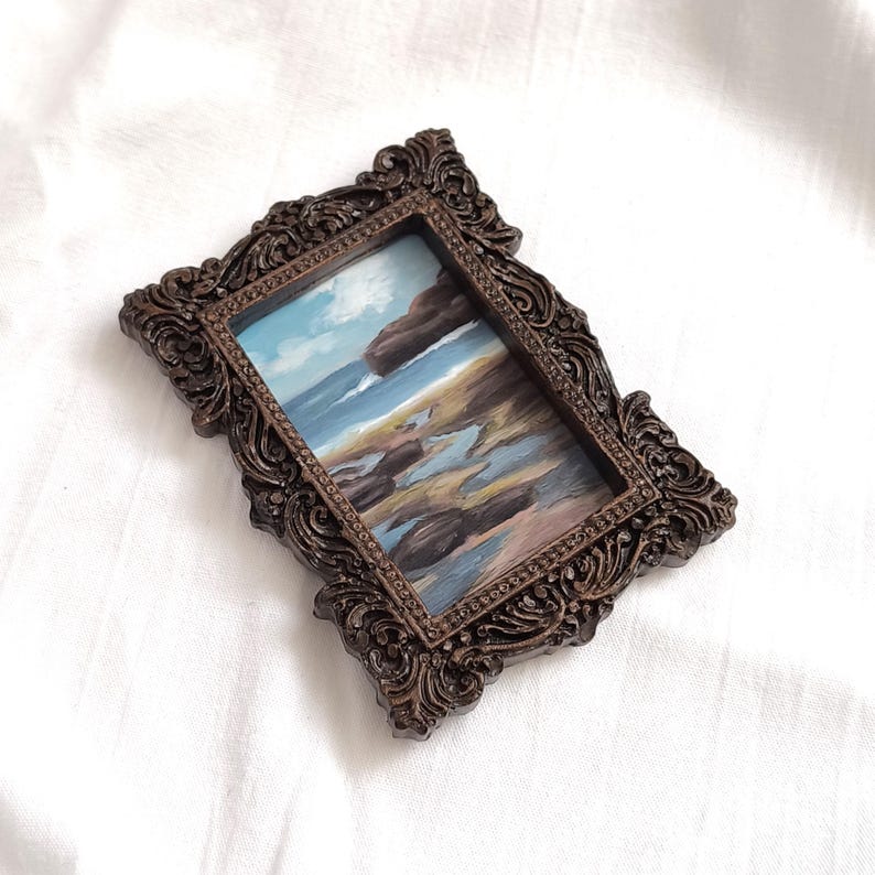 Op de afbeelding: Een rechthoekig schilderij van een kustlandschap met een blauwe oceaan, rotsachtige kustlijn en een bewolkte lucht, gevat in een sierlijke, donkerbruine, uitvoerig gesneden lijst met krulwerkdetails en een kralenrand aan de binnenzijde.