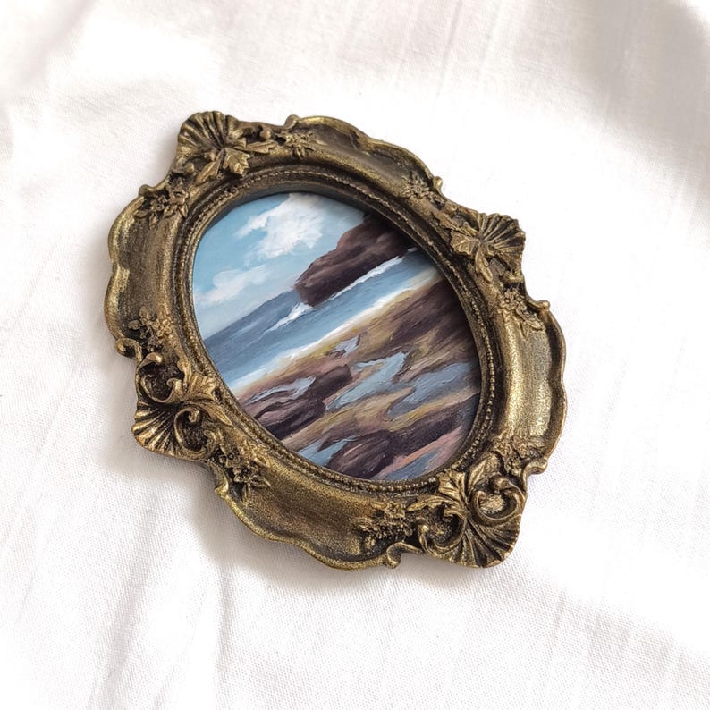 Op de afbeelding: Een ovaal schilderij van een kustlandschap met donkere rotsen, een zandstrand en een blauwe lucht met witte wolken, gevat in een sierlijke, antiek-goudkleurige lijst met gedetailleerde reli&euml;fsnijwerken langs de randen.