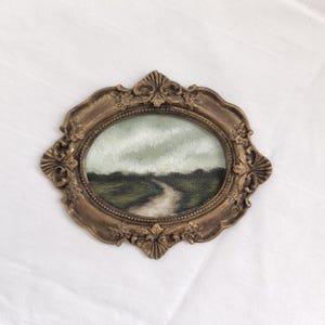 Pode incluir: Pequena pintura oval em uma moldura dourada vintage. A obra retrata uma paisagem com um caminho sinuoso através de campos verdes sob um céu nublado. A moldura tem detalhes ornamentais.