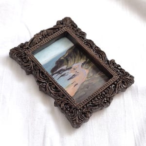 May include: A rectangular miniature painting featuring a coastal landscape with cliffs and a sandy beach, encased in a dark brown ornate frame with intricate scrollwork and floral relief details along the edges, presented in a portrait orientation.