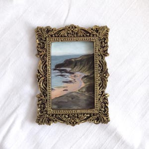 May include: A rectangular painting of a coastal landscape featuring a sandy beach, dark cliffs, and blue water, encased in an ornate, metallic gold-colored frame with intricate scrollwork and a beaded inner border detail.