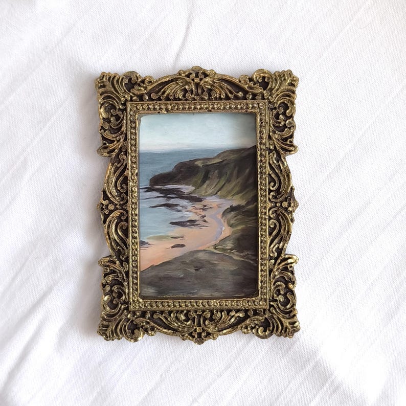 May include: A rectangular painting of a coastal landscape featuring a sandy beach, dark cliffs, and blue water, encased in an ornate, metallic gold-colored frame with intricate scrollwork and a beaded inner border detail.