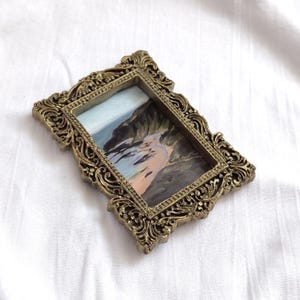 May include: A small rectangular painting of a coastal landscape featuring dark cliffs and a sandy beach, encased in an ornate, antique-style gold-colored frame with intricate scrollwork and raised relief detailing along the edges.