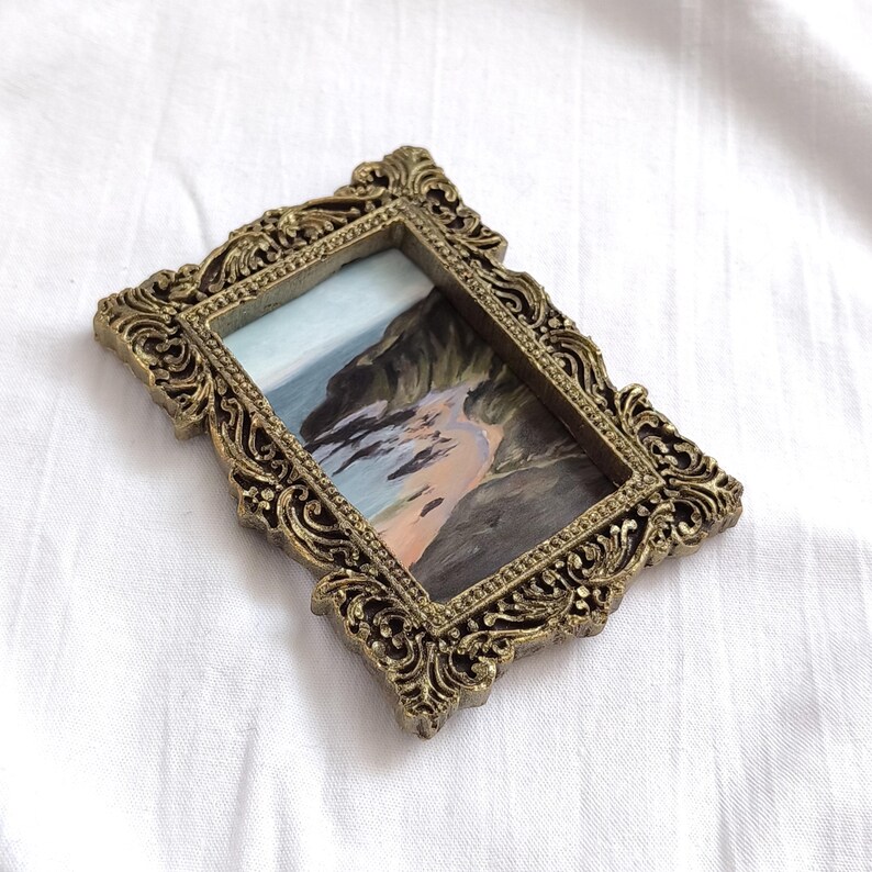 May include: A small rectangular painting of a coastal landscape featuring dark cliffs and a sandy beach, encased in an ornate, antique-style gold-colored frame with intricate scrollwork and raised relief detailing along the edges.