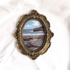 Op de afbeelding: Een ovaal schilderij van een kustlandschap met een rotsachtige kustlijn, blauwe oceaangolven en een bewolkte lucht, gevat in een sierlijke, antiek ogende goudkleurige lijst met gedetailleerde bloem- en schelpvormige reli&euml;fversieringen.