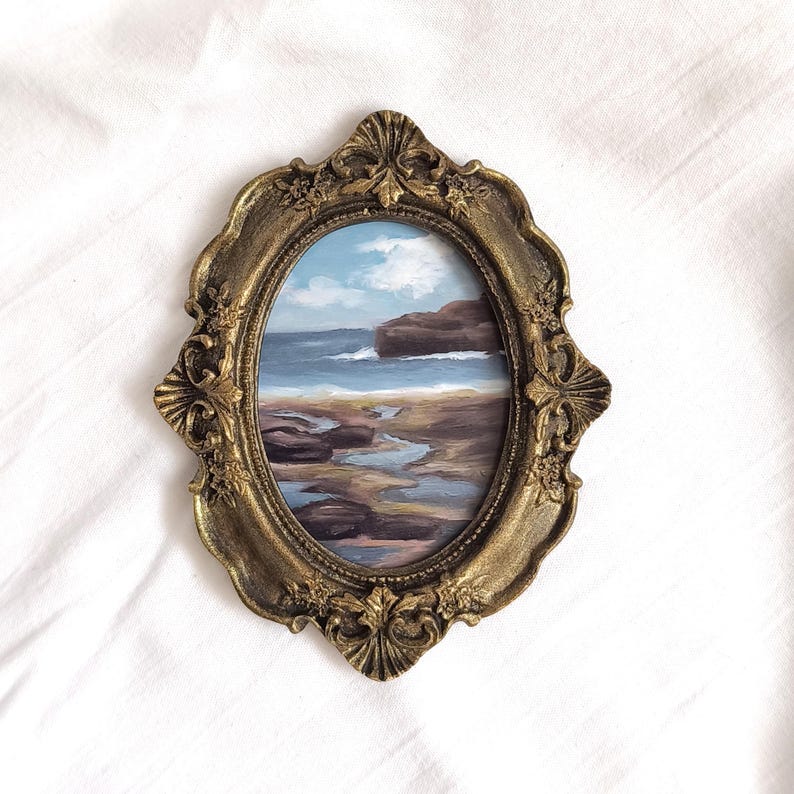Op de afbeelding: Een ovaal schilderij van een kustlandschap met een rotsachtige kustlijn, blauwe oceaangolven en een bewolkte lucht, gevat in een sierlijke, antiek ogende goudkleurige lijst met gedetailleerde bloem- en schelpvormige reli&euml;fversieringen.