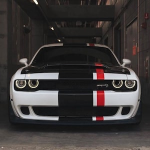 May include: A white sports car with black and red racing stripes. The front of the car features a black grille and the text "SRT". The car is parked in a garage.