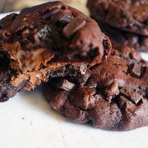 May include: Close-up of several dark chocolate cookies, one broken open to reveal a creamy chocolate filling. The cookies are studded with chocolate chunks, and the image is set against a white background. A delicious treat.