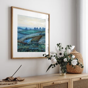 Op de afbeelding: Een ingelijst aquarel schilderij van een landschap met een blauwe lucht, groene velden en een paar bomen in de verte. Het schilderij hangt aan een witte muur boven een houten dressoir met een vaas met witte bloemen en een rieten mand.