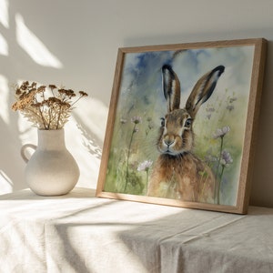 May include: Watercolour painting of a brown hare with large ears, looking directly at the viewer. The hare is surrounded by a field of green grass and small pink flowers.
