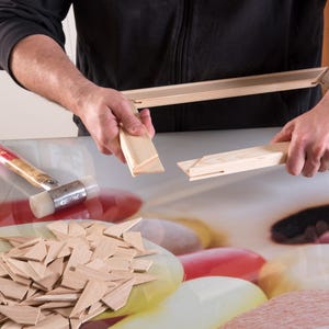 Può includere: Una persona sta assemblando una cornice di legno per un dipinto su tela. La cornice è fatta di sottili strisce di legno e viene tenuta insieme con un martello di gomma. Ci sono anche un mucchio di cunei di legno e un mucchio di strisce di legno.