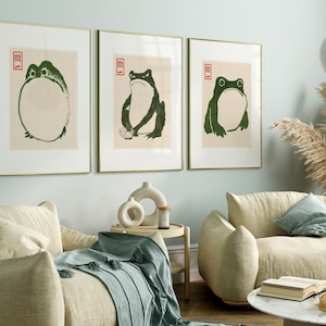 May include: Three framed prints of frogs in a minimalist style. The prints are in gold frames and are hung on a light blue wall. The frogs are depicted in black and green.