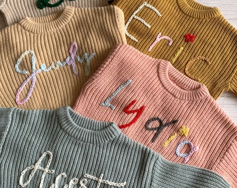May include: Four knitted sweaters with different names embroidered on them. The sweaters are in various colors, including beige, mustard yellow, pink, and green. The names are "Darcey", "Eric", "Lily", and "Ace"t.