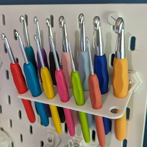 May include: A set of 12 colorful crochet hooks, organized in a white plastic holder. The hooks are arranged in a row, with the handles facing up. The hooks are made of metal and have a variety of colors, including red, blue, yellow, green, and pink.
