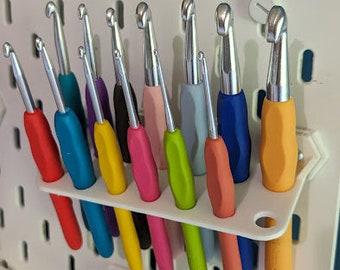 Pode incluir: Um conjunto de 12 agulhas de crochê coloridas, organizadas em um suporte de plástico branco. As agulhas estão dispostas em uma fileira, com as alças voltadas para cima. As agulhas são feitas de metal e têm uma variedade de cores, incluindo vermelho, azul, amarelo, verde e rosa.