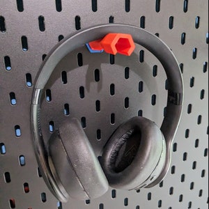 May include: Black over-ear headphones with a black headband hanging on a pegboard wall. The headphones are held in place by a red and blue 3D printed hook.