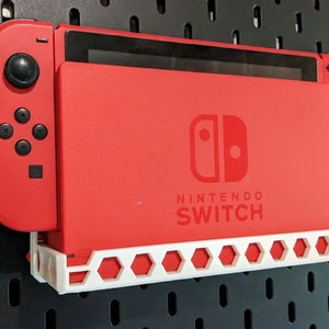 May include: A red Nintendo Switch console with a white wall mount. The console is attached to the wall mount with a white plastic bracket. The console is in a closed position, with the screen facing the wall.