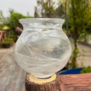 Puede incluir: Un jarrón de vidrio transparente con forma redondeada y superficie texturizada. El jarrón está colocado sobre un poste de madera.