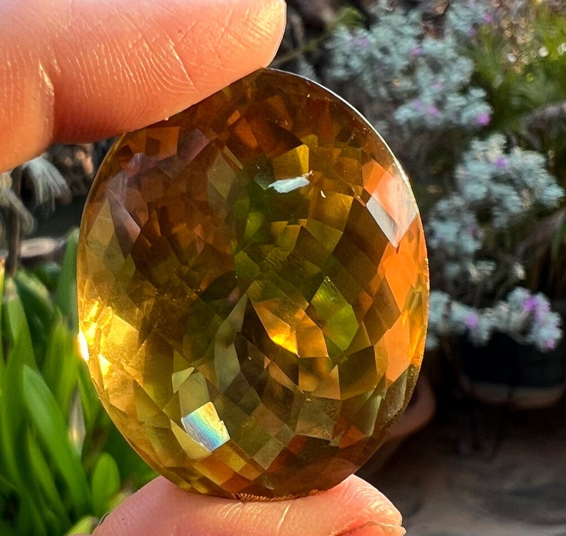 May include: A large, faceted, oval-shaped gemstone with a golden yellow color and green and iridescent hues.