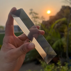 以下が含まれることがあります： 夕焼けと葉のぼやけた背景を背景に、手の中に保持された長方形の透明なガラスプリズム。