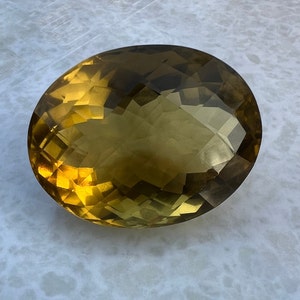 May include: A large, oval-shaped, faceted gemstone with a golden yellow color and a slight green hue.