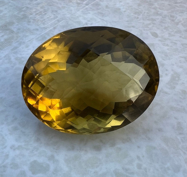May include: A large, oval-shaped, faceted gemstone with a golden yellow color and a slight green hue.