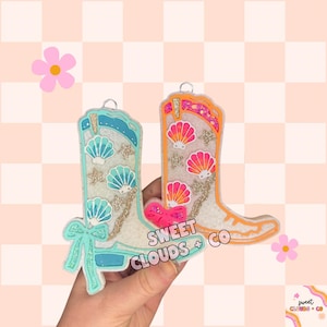 May include: Two decorative cowboy boot ornaments. One is light blue with seashell and star accents, the other is orange with seashell and star accents. Both have glitter details and the text "SWEET CLOUDS + CO".