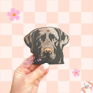 May include: A wooden cutout of a chocolate Labrador Retriever dog head. The cutout is painted black and brown and has a distressed wood look. The cutout is held in a hand and the text "SWEET CLOUDS + CO" is visible in the bottom left corner of the image.