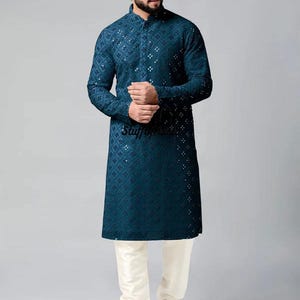 Puede incluir: Una kurta de manga larga azul turquesa con un patrón geométrico blanco. La kurta tiene un cuello mandarín y un cierre de botón. El modelo lleva pantalones blancos y zapatos blancos.