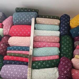 Pu&ograve; includere: Esposizione di vari rotoli di tessuto in una variet&agrave; di colori, tra cui rosso, verde, blu e giallo. Ogni rotolo &egrave; adornato con un motivo ripetuto di piccoli abbellimenti riflettenti, creando un aspetto strutturato. I tessuti sono ordinatamente impilati sugli scaffali.
