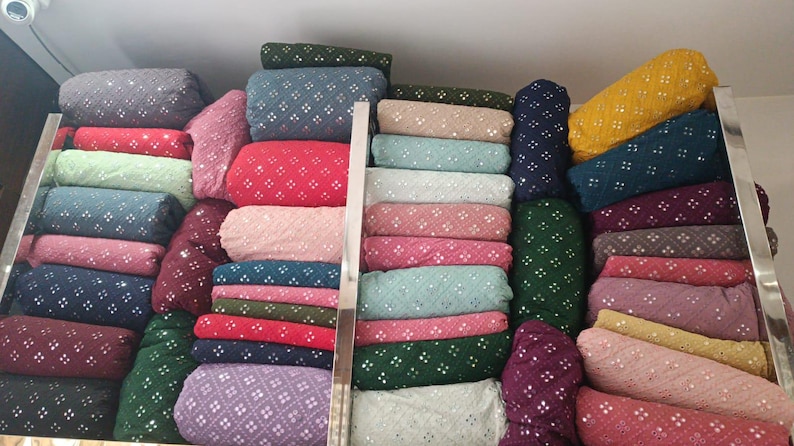 Pu&ograve; includere: Esposizione di vari rotoli di tessuto in una variet&agrave; di colori, tra cui rosso, verde, blu e giallo. Ogni rotolo &egrave; adornato con un motivo ripetuto di piccoli abbellimenti riflettenti, creando un aspetto strutturato. I tessuti sono ordinatamente impilati sugli scaffali.