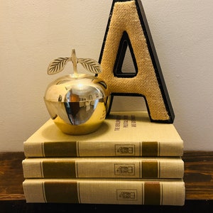 May include: A silver apple shaped trinket box sits on top of a stack of four books. The books have a beige linen cover with a brown stripe and a small gold design. A large burlap letter A leans against the stack of books.